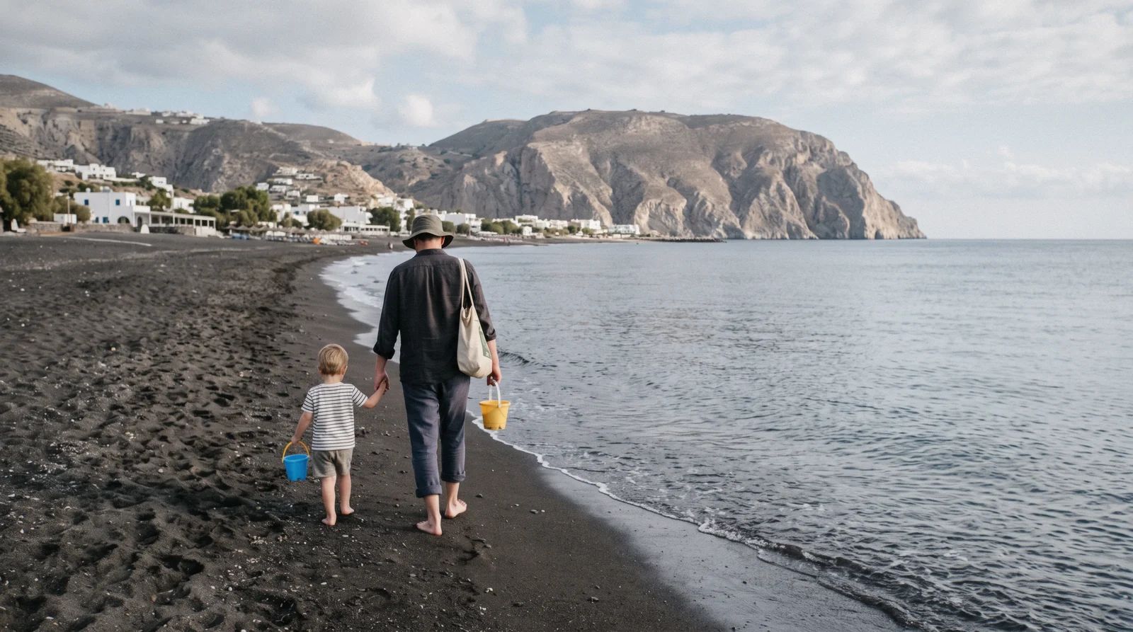A parent and a child seen from behind walking hand in hand along the black sand at Kamari in the morning, santorini with kids, Santorini Greece